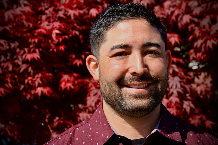 A smiling person stands in front of a maple tree who's leaves have all turned red
