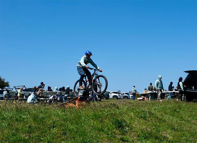 A person jumps a bike onto some grass. A number of people walk around in the background