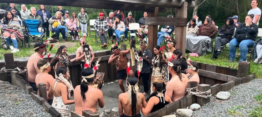 A group of smiling young adults in traditional dress perform a ceremony while people watch on