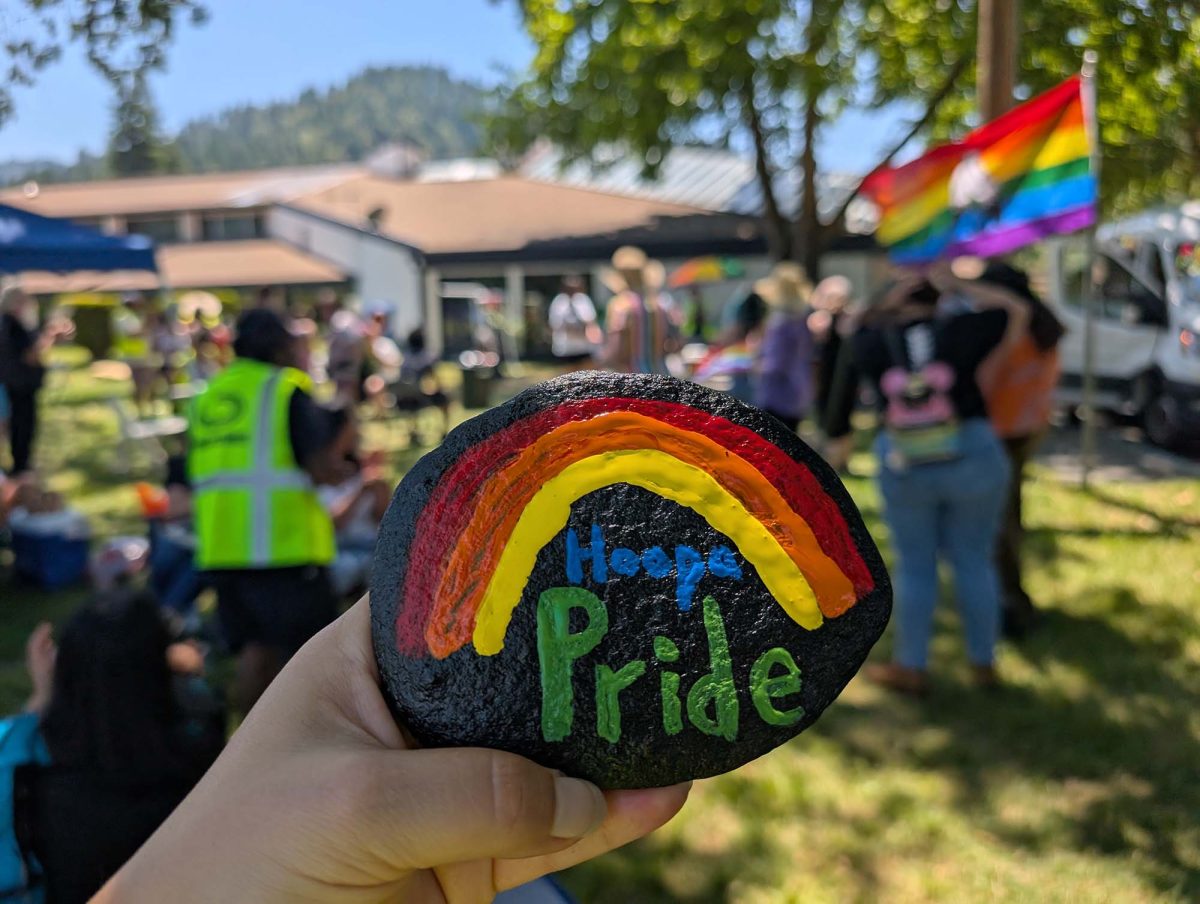 A hand holds up a rock painted with a rainbow and "Hoopa Pride". In the background is a community event with rainbow flags and a band.