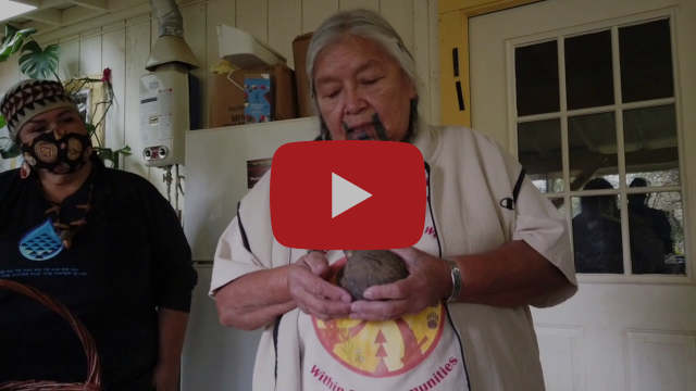 A Native American elder holds an item, in a screenshot from a video. A large YouTube play button sits over the middle of the image