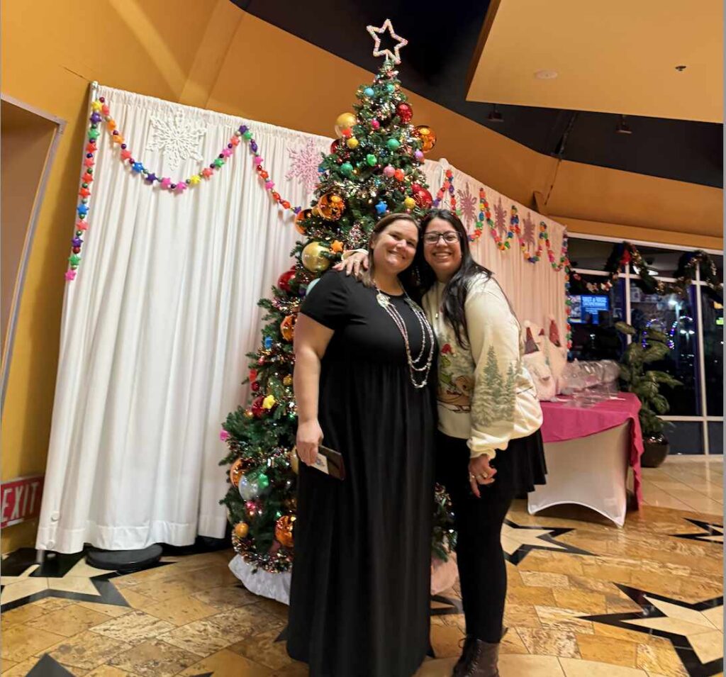 Two women hug and pose for the camera. The room in the background is well-decorated for a holiday party