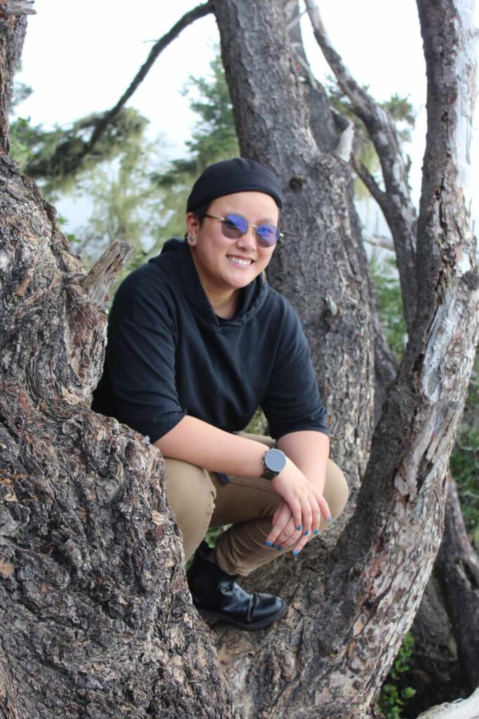 A smiling young adult stands in a tree and poses for the camera
