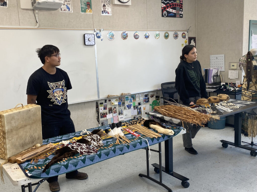 Two people stand in a classroom and give a presentation. In front of them are two long tables full of traditional Native American items