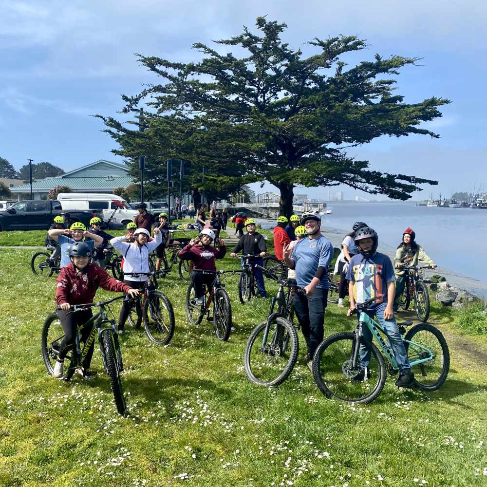 A group of people pose for the camera on their bikes. In the background is a large tree and the bayfront