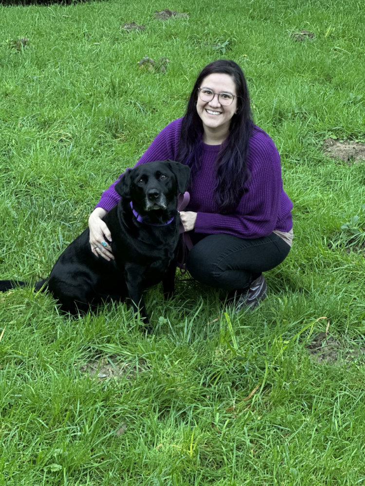 A smiling person crouches in a field and pets a dog
