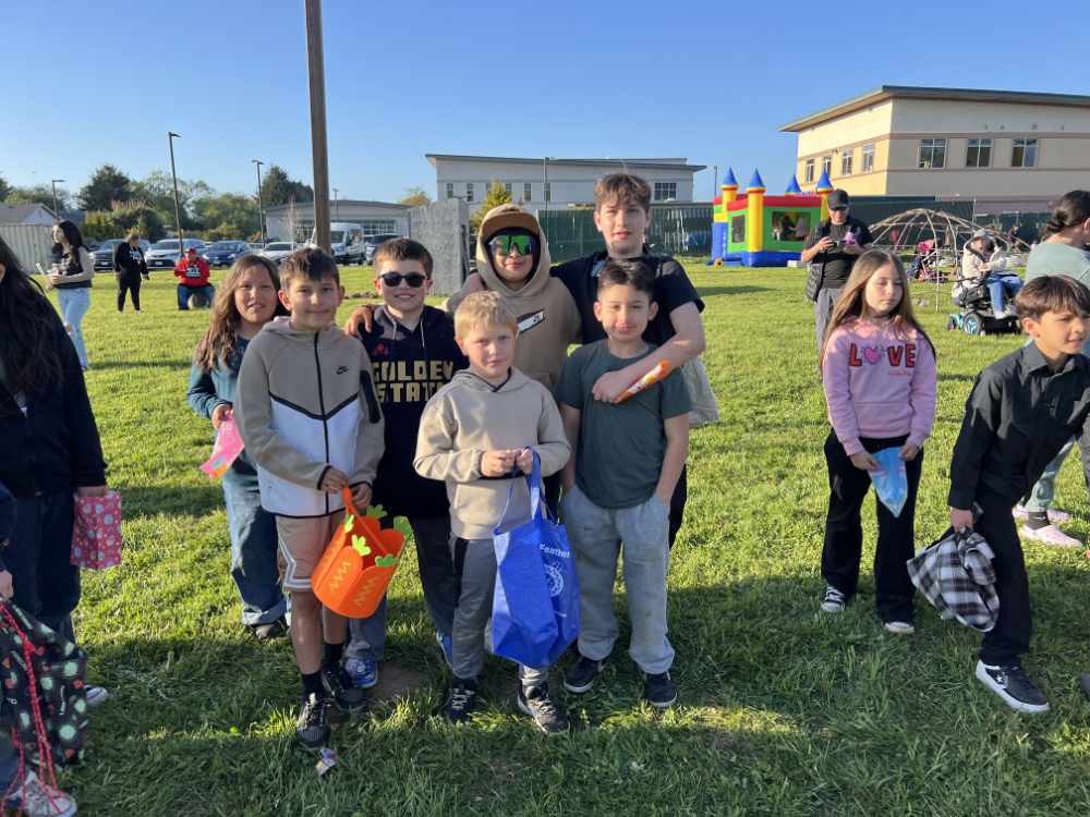 A group of youth pose together in a park. In the background is a festival with some inflatable bounce houses