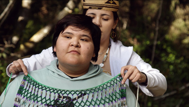 A Native American woman has a traditional beaded item draped over her by another Native woman
