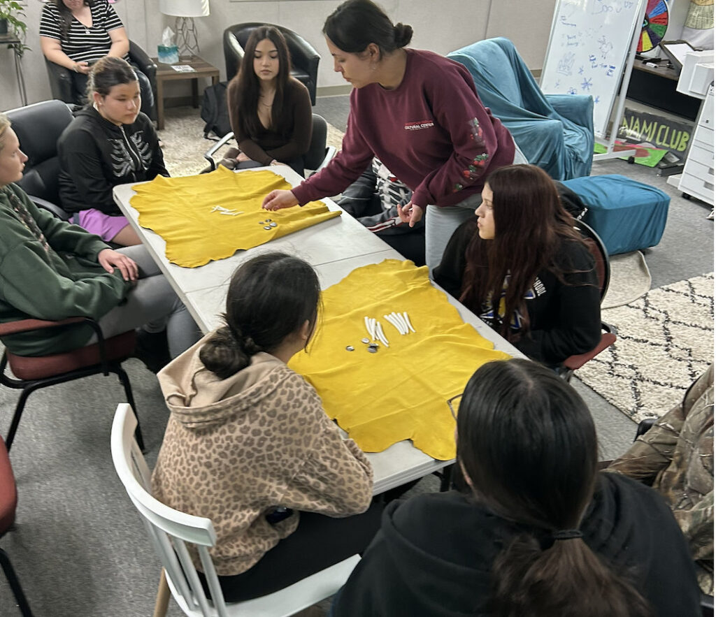 Several young adults sit around a table and watch as an adult demonstrates shell dice