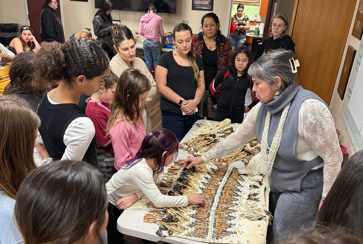 An adult stands behind a table and demonstrates traditional native dress to a group of younger people