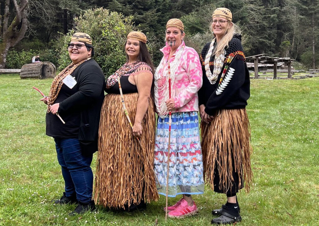 Four Native American women in traditional dress stand in a meadow and pose for the camera